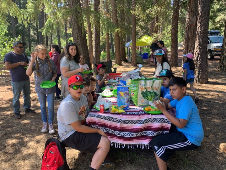 Families sitting at an outdoor picnic table with a Mexican blanket tablecloth eating lunch on a sunny day with the forest in the background.