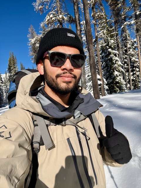 BIPOC male in winter clothing and beanie in the snow giving a thumbs up