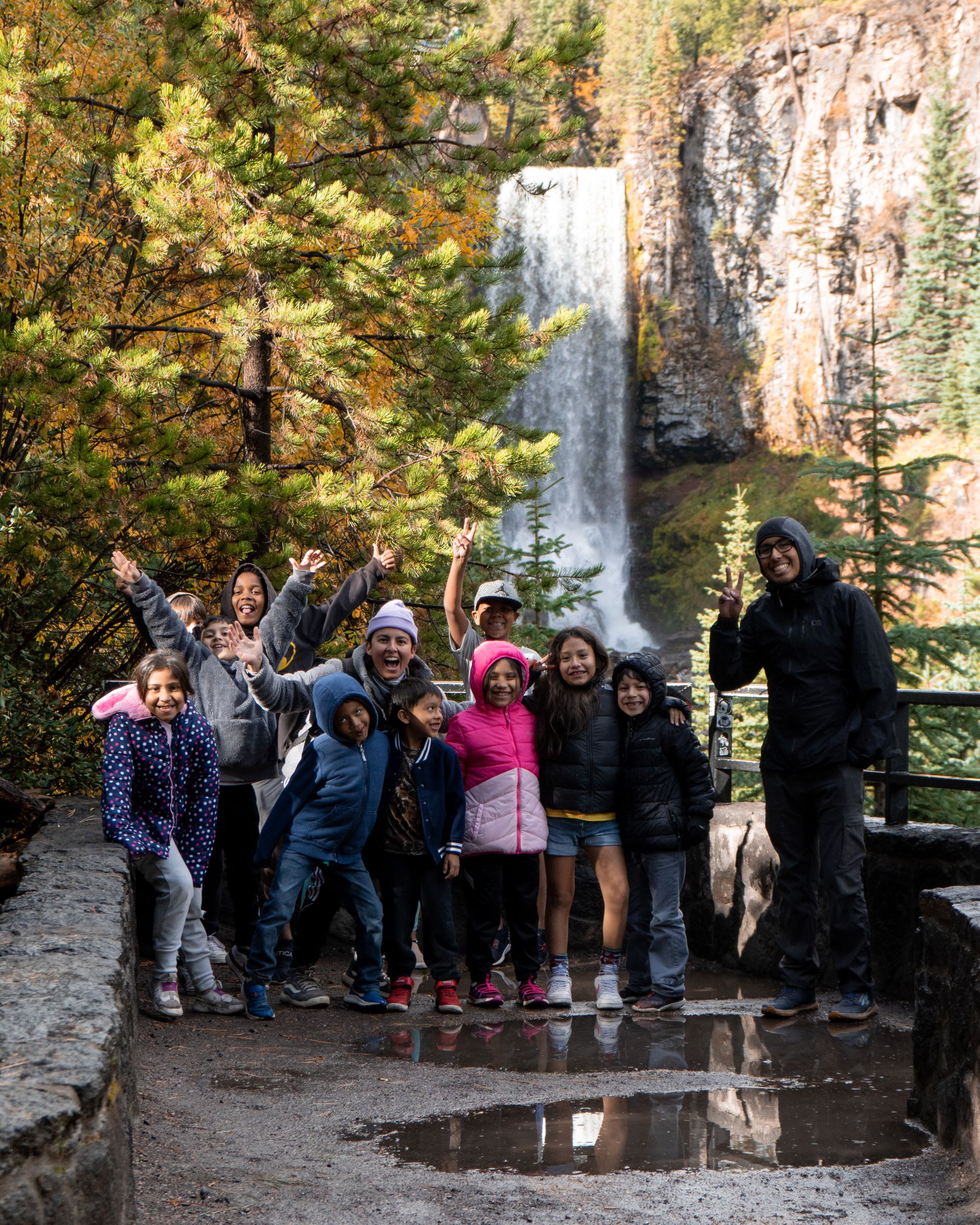 A group of 10 latino youth stand in front of a waterfall smiling with arms in the air with 2 Vamonos Outside group leaders